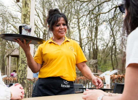 Medewerker van Landal serveert drankjes aan gasten op een terras in een bosrijke omgeving.